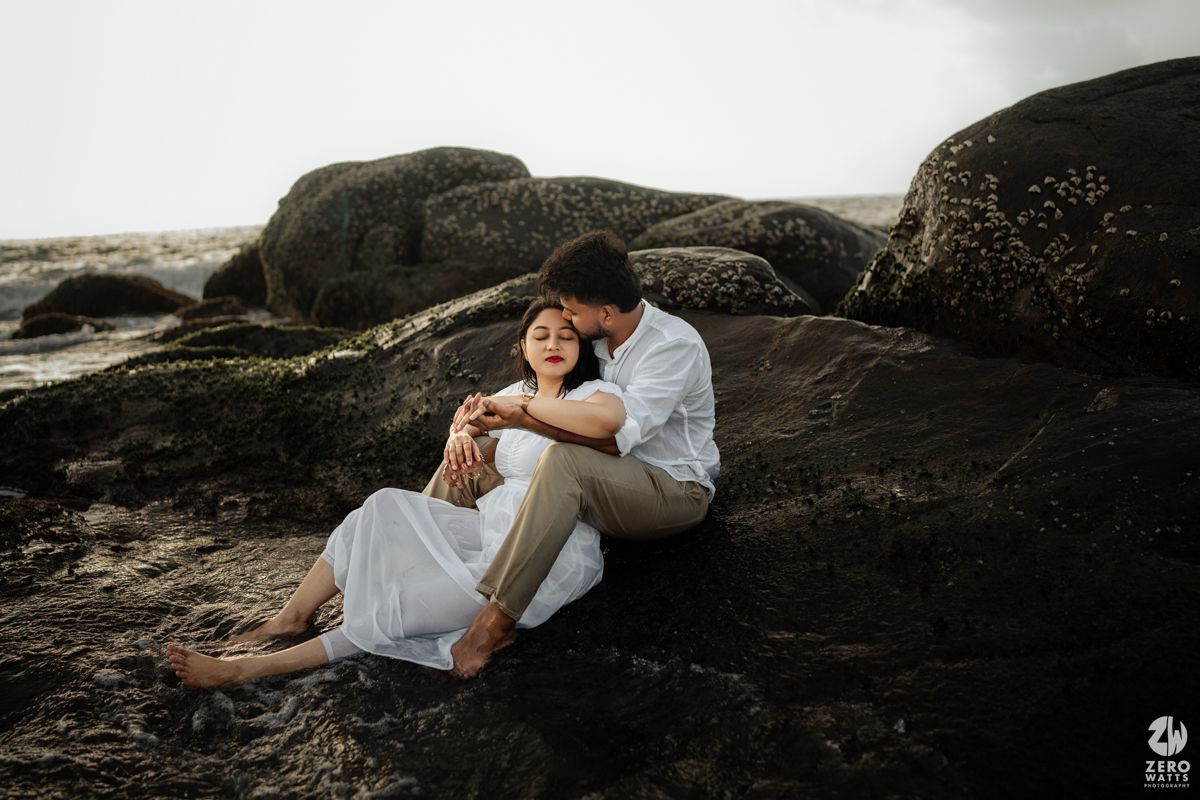 Couple in white clothing sharing intimate moment while sitting on coastal rocks at sunset.