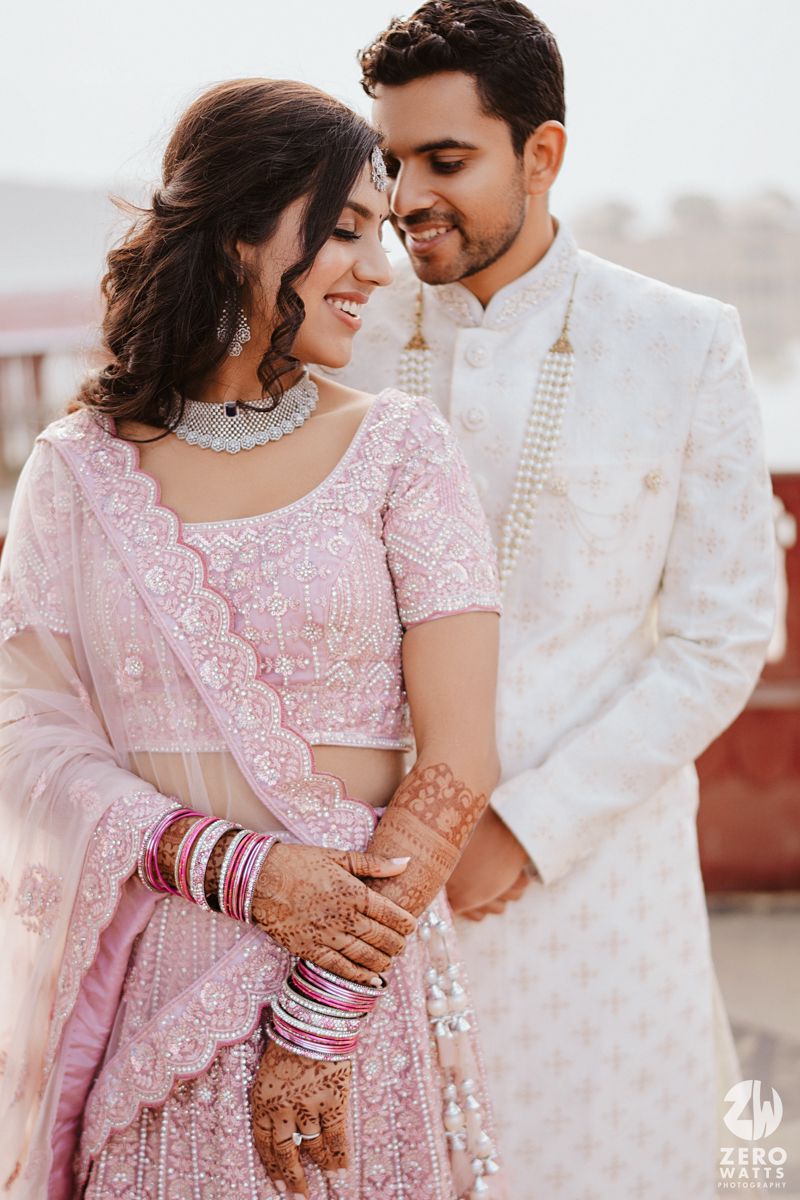 A romantic couple in traditional Indian wedding attire share an intimate moment wearing pink lehenga and white sherwani.