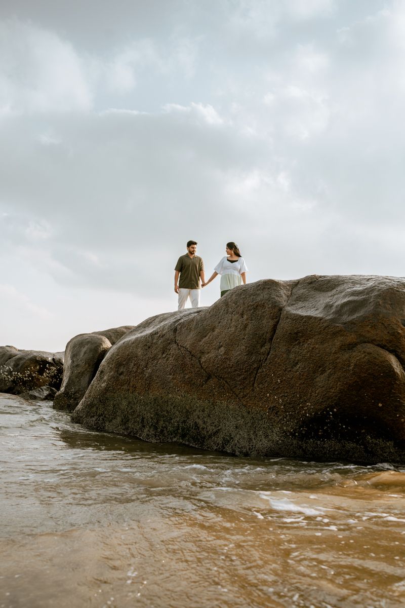 Couple standing together on large coastal rocks with cloudy sky and ocean waves below.