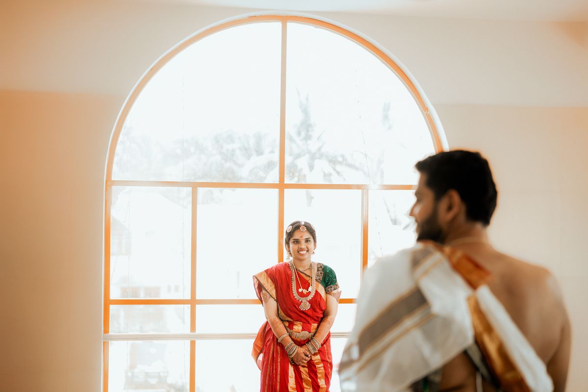 A person in a vibrant red saree stands by an arched window while someone in white is seen in the foreground.