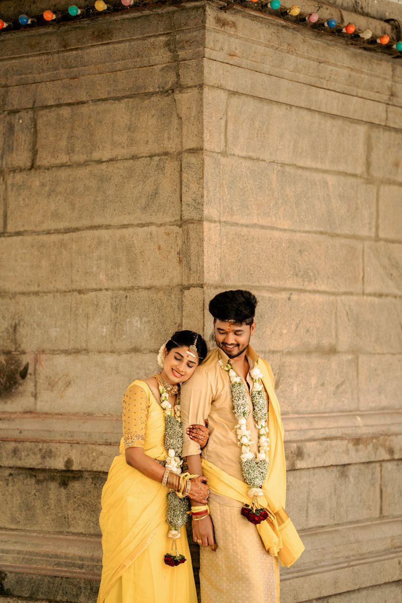 A traditional South Indian wedding couple in matching yellow attire pose together against a stone wall with string lights.