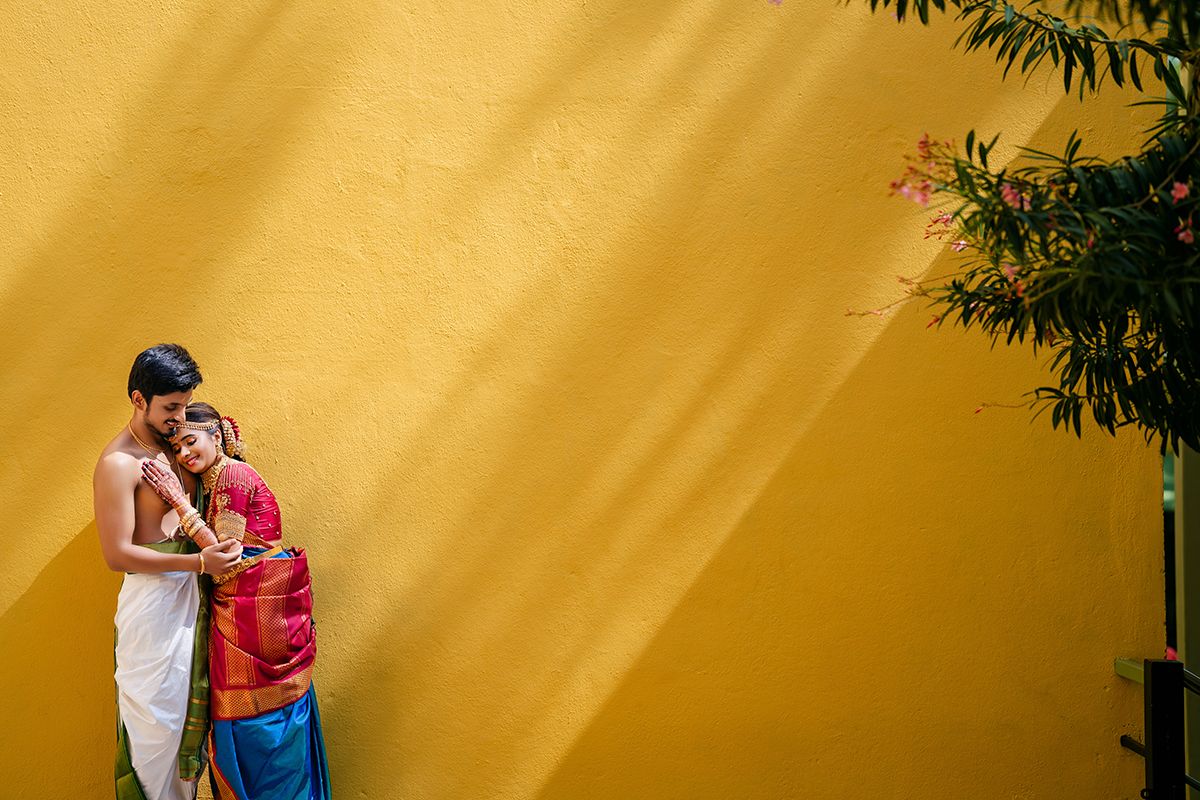 Two people stand close together against a vibrant yellow wall with dramatic diagonal sunlight and bamboo plants.