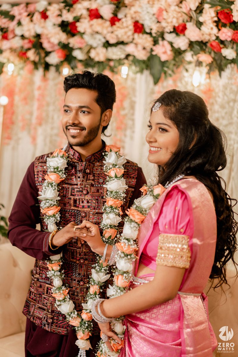A couple in traditional Indian wedding attire with floral garlands stands against a decorated backdrop with pink flowers.