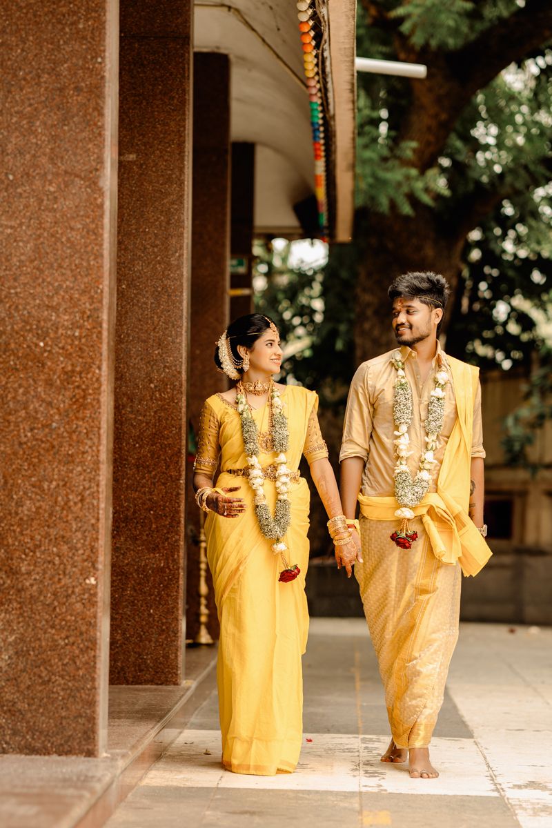 A couple wearing matching yellow traditional Indian outfits with floral garlands walk together in a covered walkway.