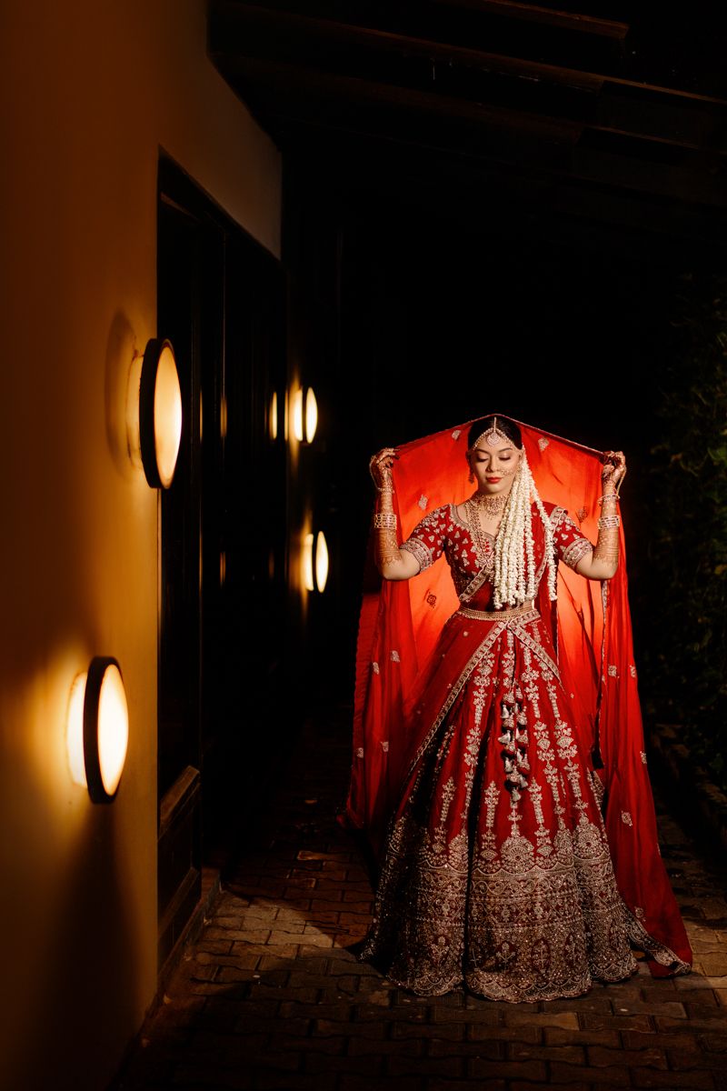 A bride in a traditional red and gold embellished Indian lehenga stands elegantly against a warmly lit wall with circular sconces.