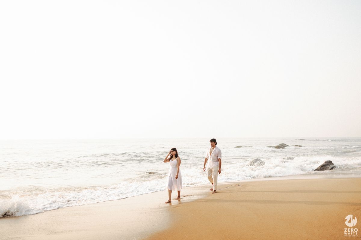 Couple in white clothing walking along a serene beach shoreline during sunset.