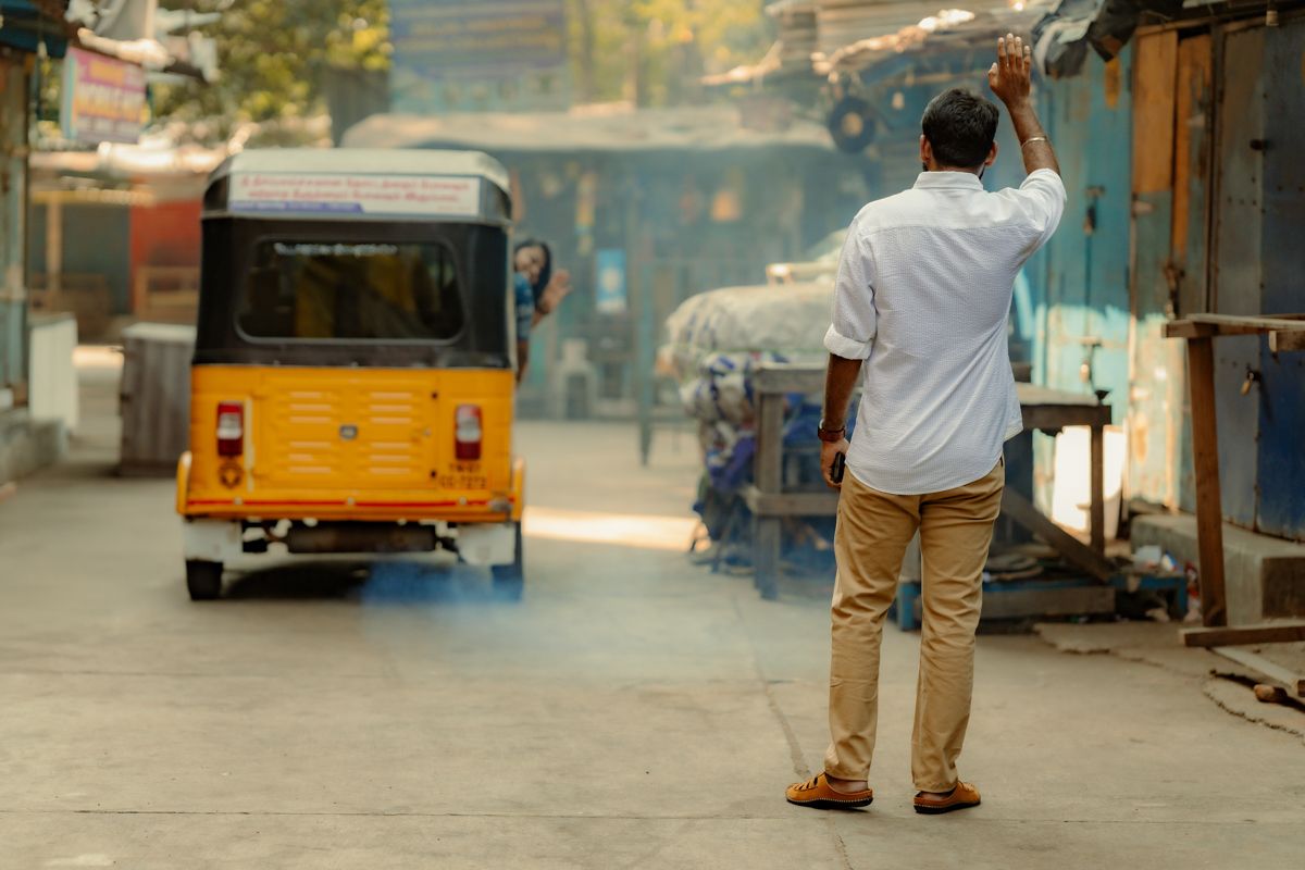 Man in white shirt stands on street facing yellow auto rickshaw in misty urban scene.