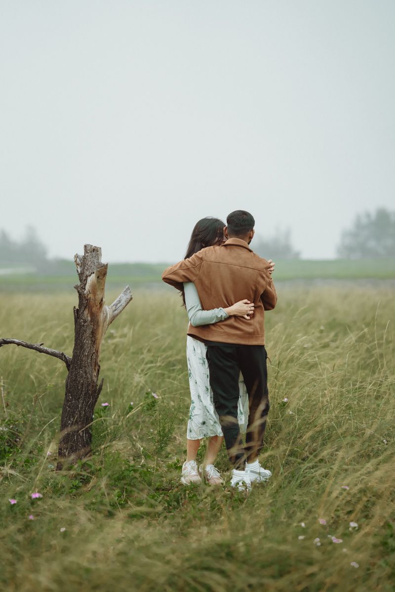 Two people embracing while standing in tall grass on a misty day, viewed from behind.