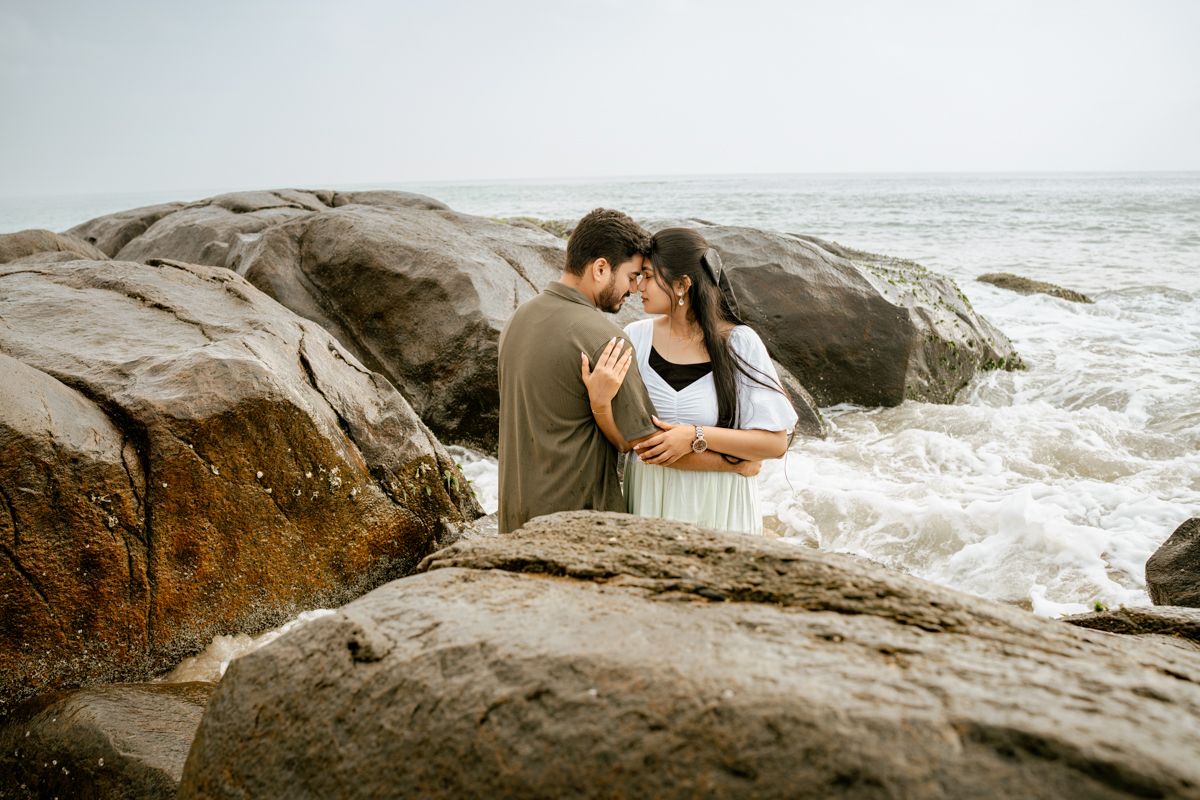 Romantic couple embracing on rocky shoreline with crashing waves in background.