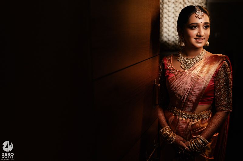 Traditional Indian bride in burgundy silk saree and ornate gold jewelry poses in dramatic low-light setting.