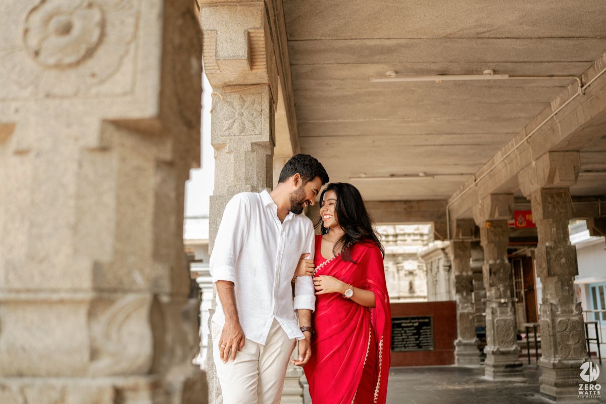 Couple in coordinated red and white outfits pose intimately along stone pillared corridor.