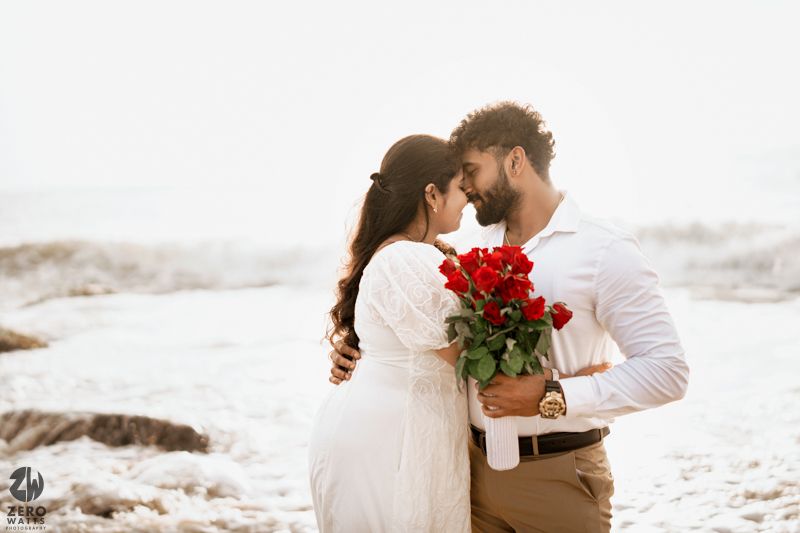 A couple shares an intimate moment on the beach while holding red roses.