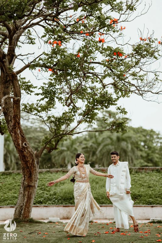 A couple dances playfully under flowering trees in a garden setting wearing elegant cream and white formal attire.
