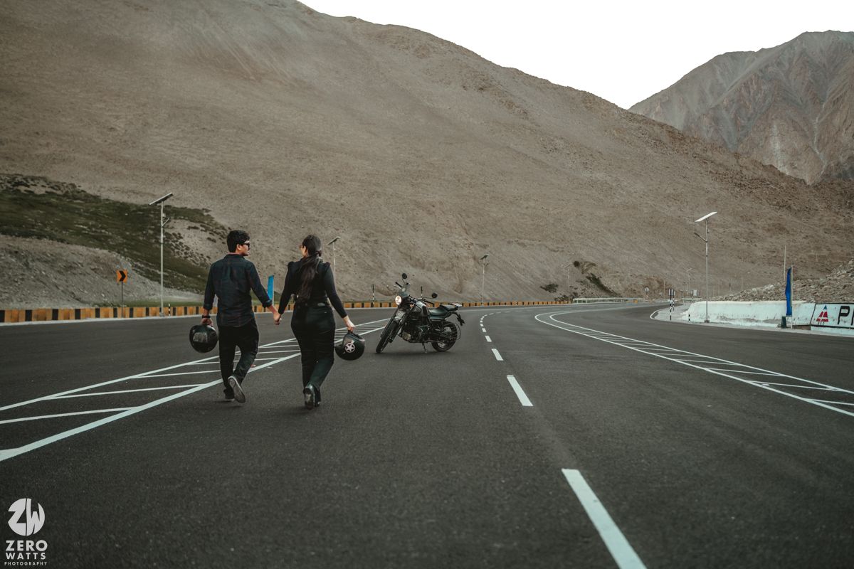 Two people walking away on a mountain road with a parked motorcycle against dramatic rocky terrain.