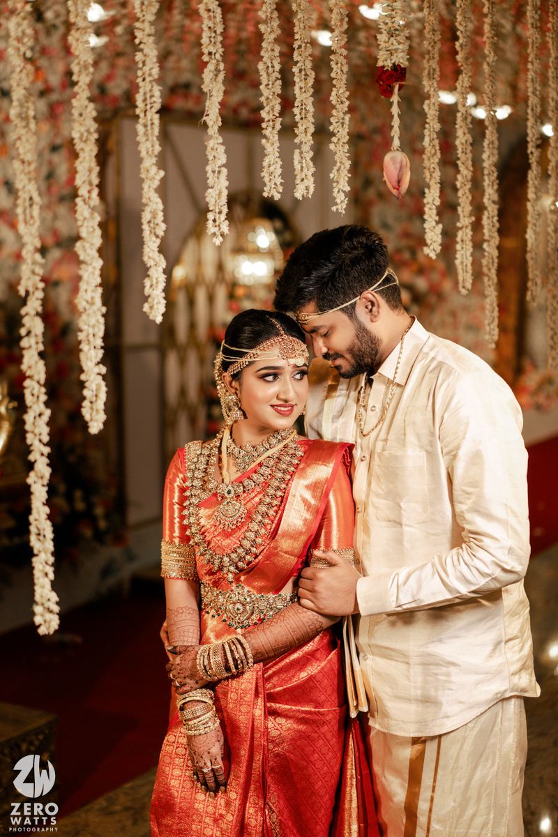 A traditional South Indian wedding couple pose beneath hanging floral decorations in elegant red and white attire.