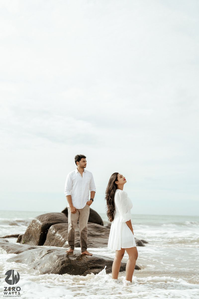 A couple in white attire stands on coastal rocks with ocean waves and overcast sky in the background.