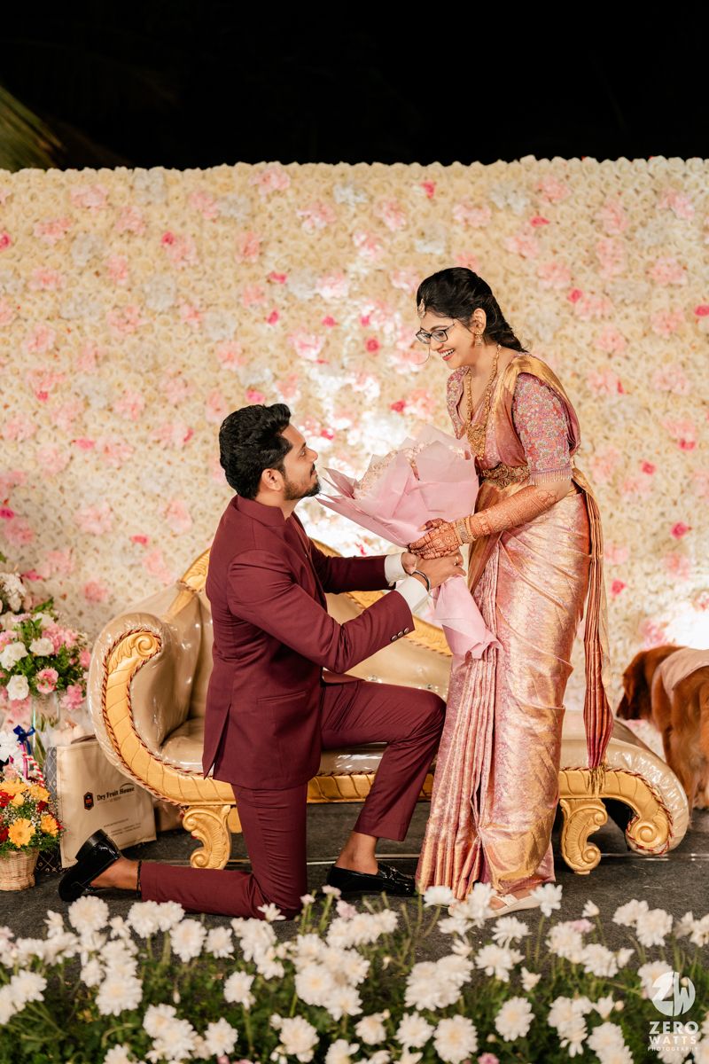 Romantic proposal scene against a floral backdrop with white flowers and pink petals scattered around.