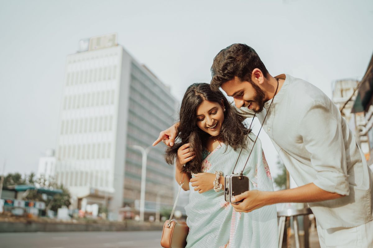 A stylishly dressed couple shares a laugh while looking at a phone on an urban street with buildings in the background.