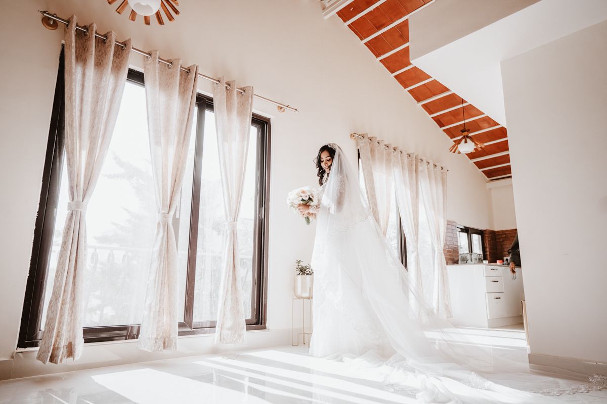 A bright bridal suite room with white curtains and exposed wooden beams features a hanging wedding dress in natural light.