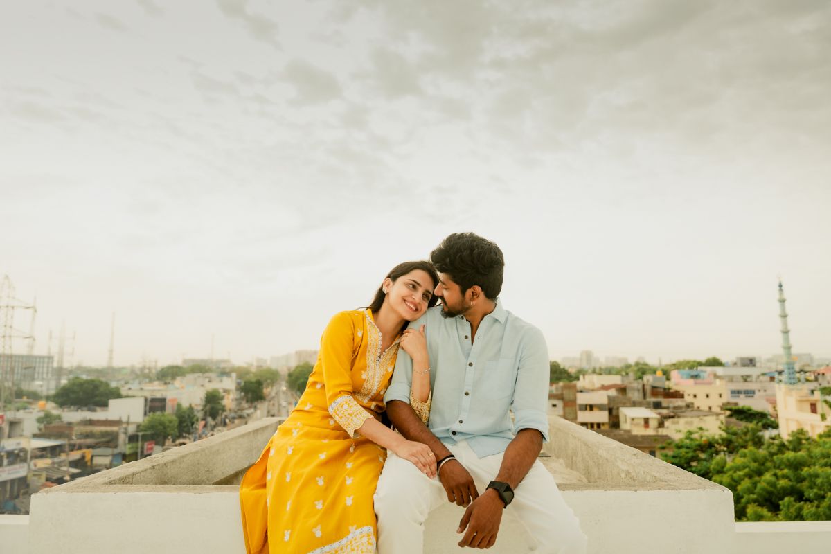 A romantic couple in yellow and white outfits share an intimate moment on a rooftop overlooking a cityscape at dusk.