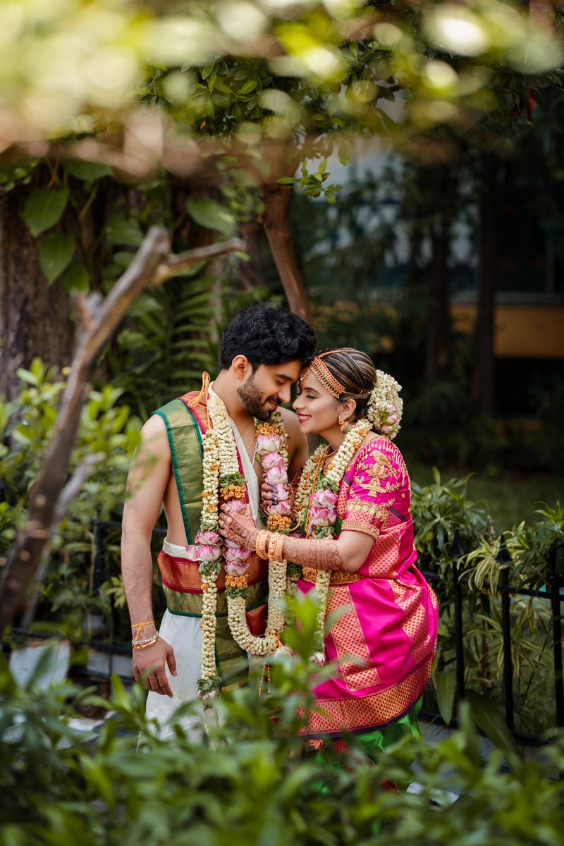 A couple in traditional South Indian wedding attire with flower garlands share an intimate moment in a garden setting.