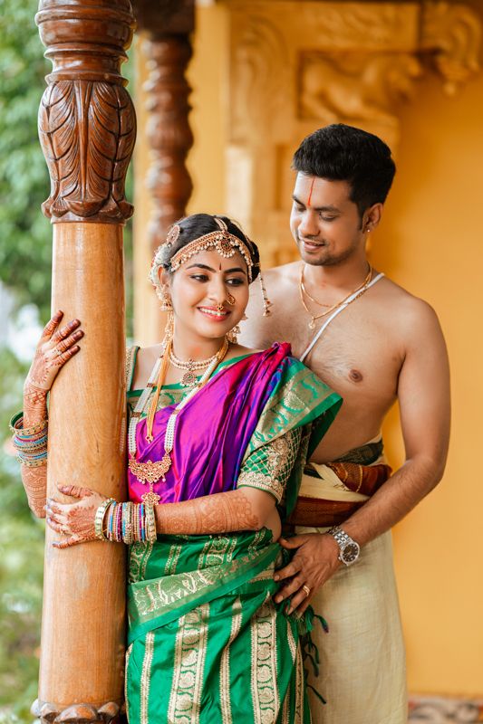 Traditional South Indian couple in wedding attire pose together against carved wooden pillars in warm lighting.