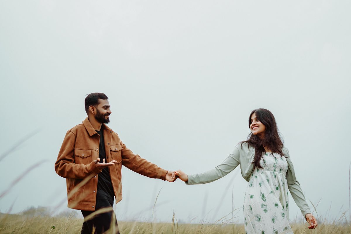 Two people hold hands while walking through a misty field, wearing brown jacket and white floral dress.