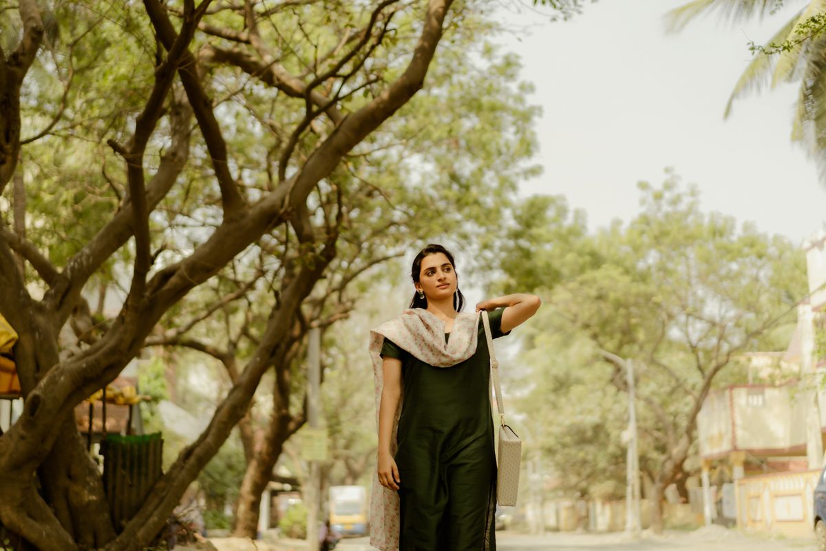 Woman in a black dress poses outdoors under lush green trees.