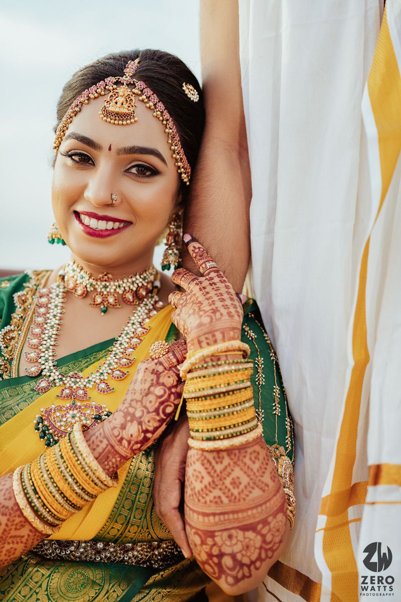 Traditional South Indian bride wearing ornate jewelry and henna adorned in yellow and green silk saree.