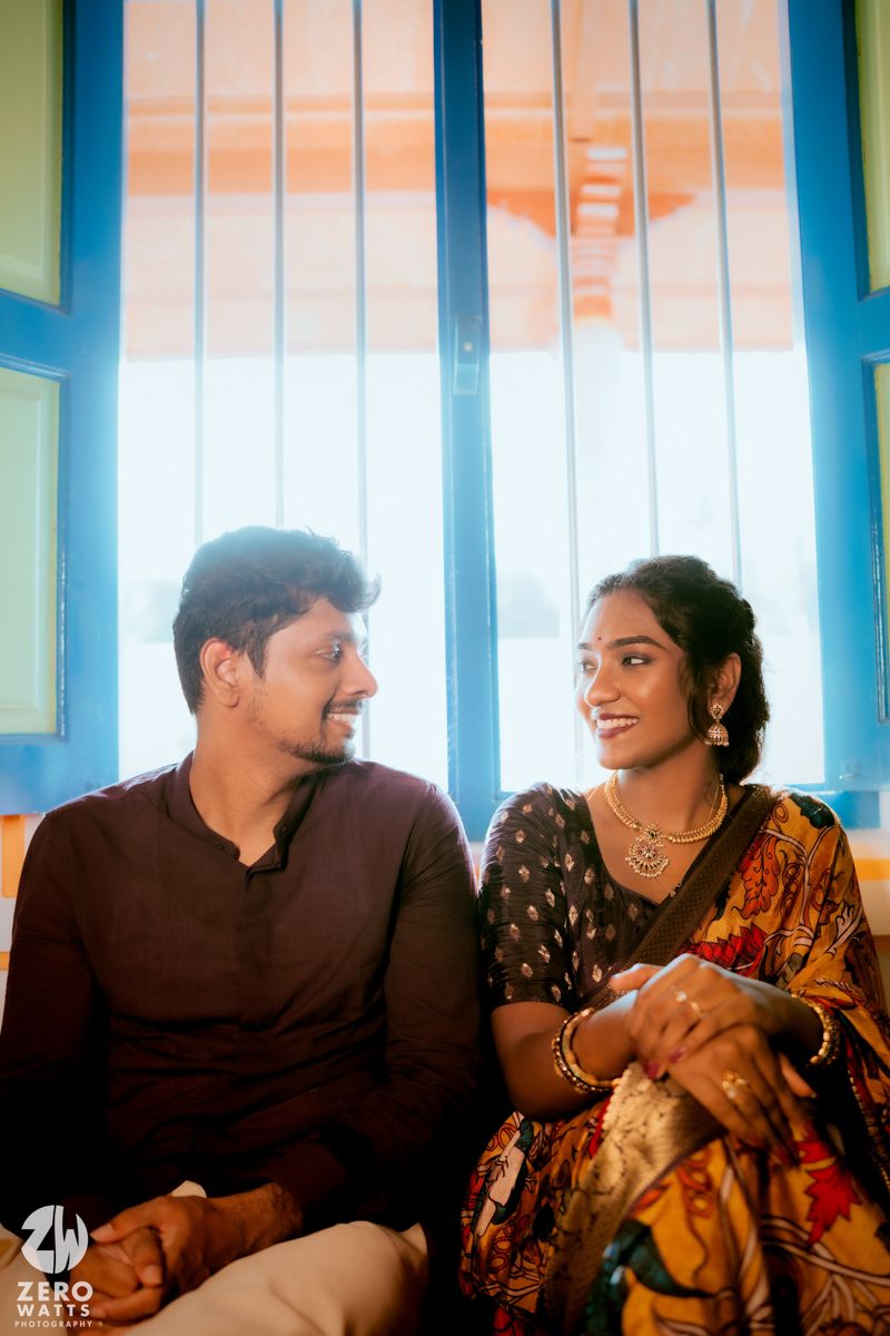 Couple in traditional Indian dress sits by window with colorful blue frames during sunset.