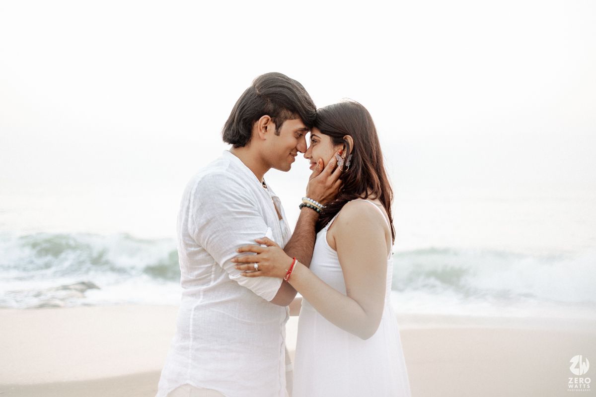 A romantic couple in white outfits share an intimate moment on a beach with ocean waves in the background.