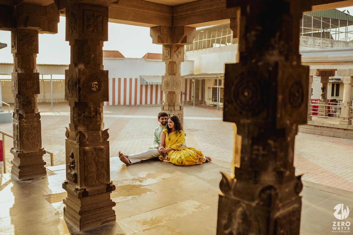 Person in yellow saree sitting on ground under carved stone pillars of a traditional temple.