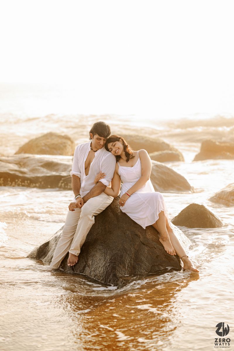 Couple in white sits together on large rocks by the ocean during golden hour.