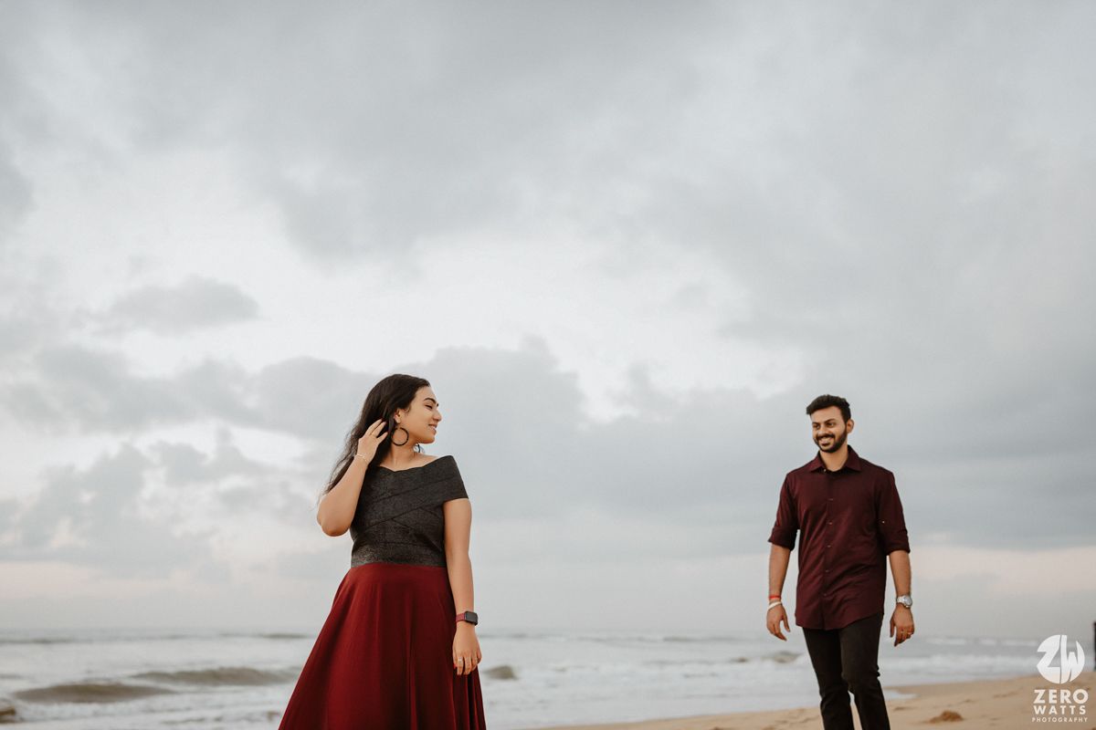 A couple stands on a beach with overcast skies, the woman wearing a red skirt while wind blows through her hair.