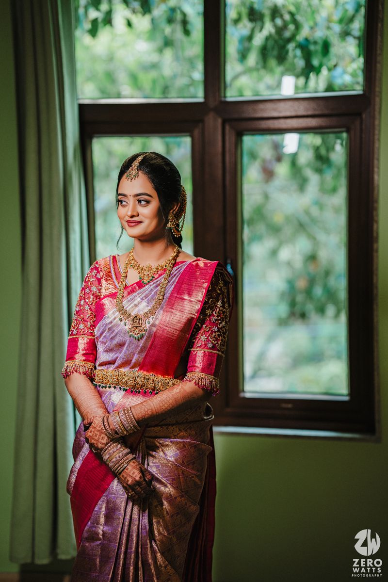 Traditional South Indian bride in a luxurious pink and purple silk saree poses elegantly by a window with greenery outside.