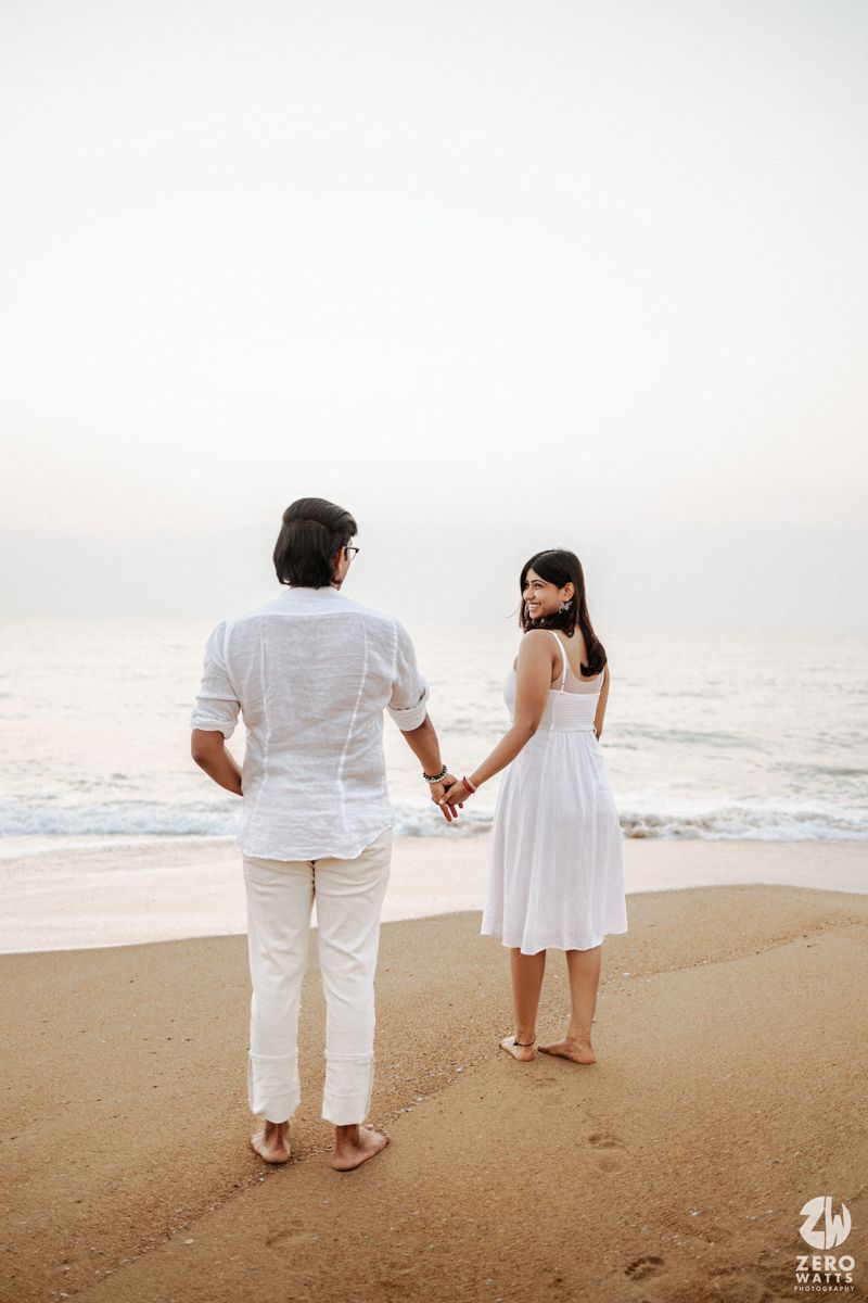 A couple in white outfits holding hands while walking along a sandy beach at sunset.