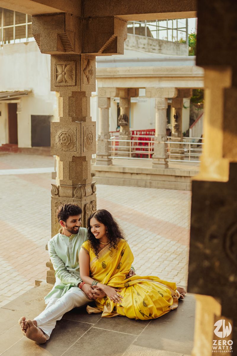 A couple in traditional Indian attire sits together under carved stone pillars in a temple courtyard.
