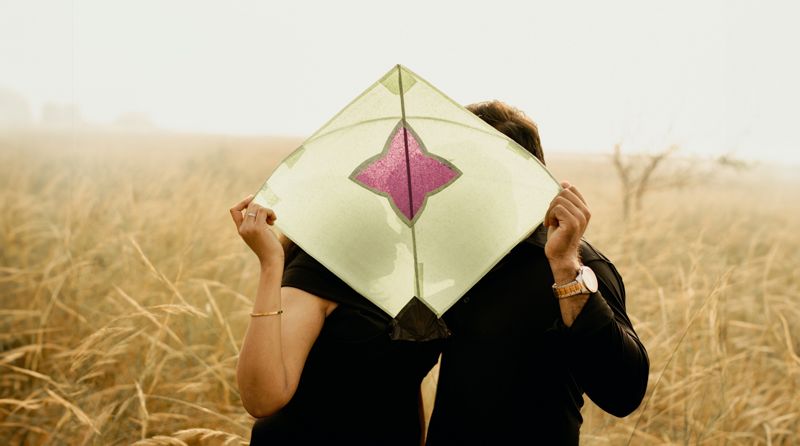 Two people holding up a diamond-shaped paper kite while standing in a golden wheat field during sunset.