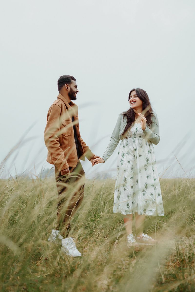 A couple holds hands while standing in a grassy meadow, wearing casual outfits on an overcast day.