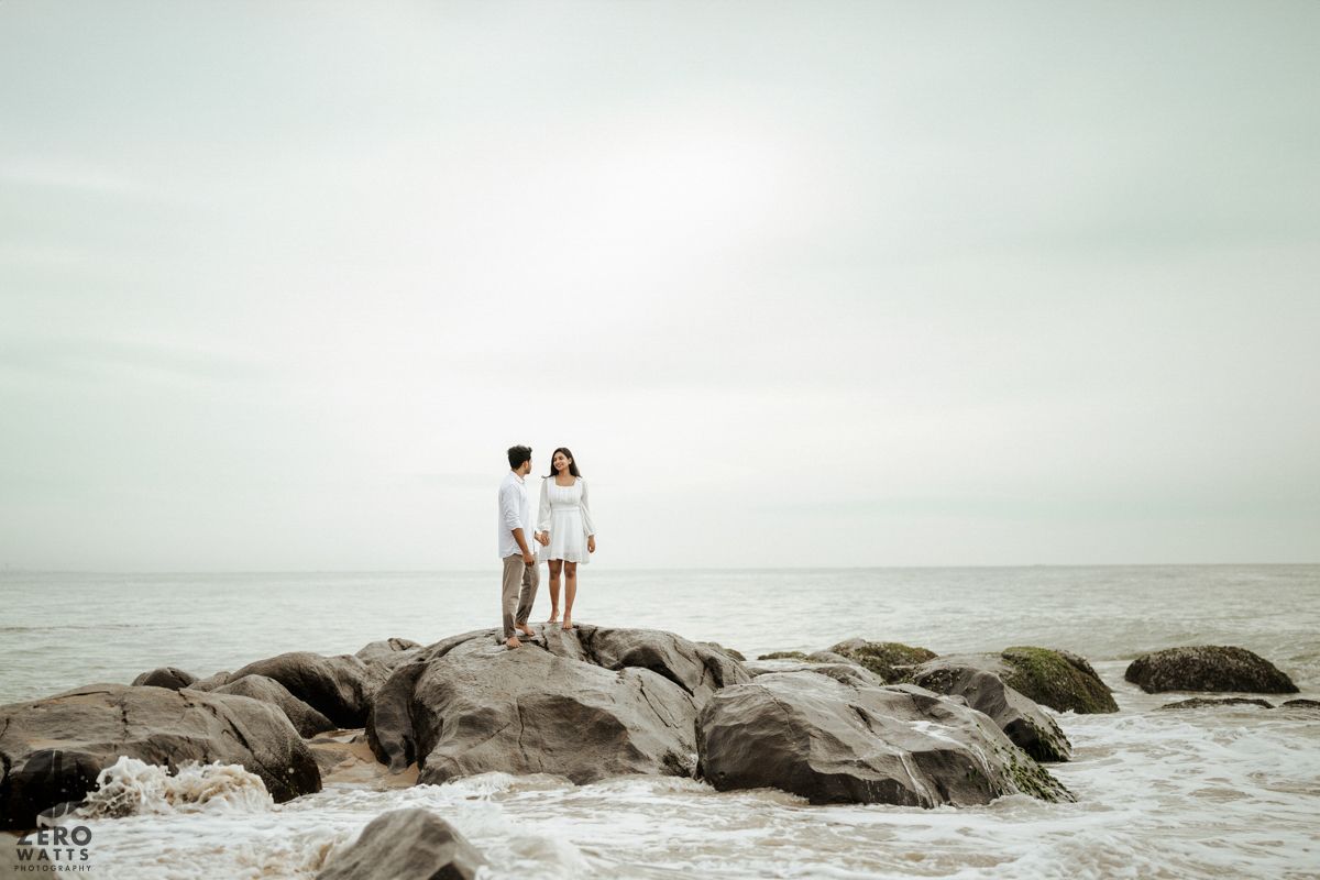 Two people stand together on rocky coastal outcrop against misty ocean and cloudy sky at dusk.