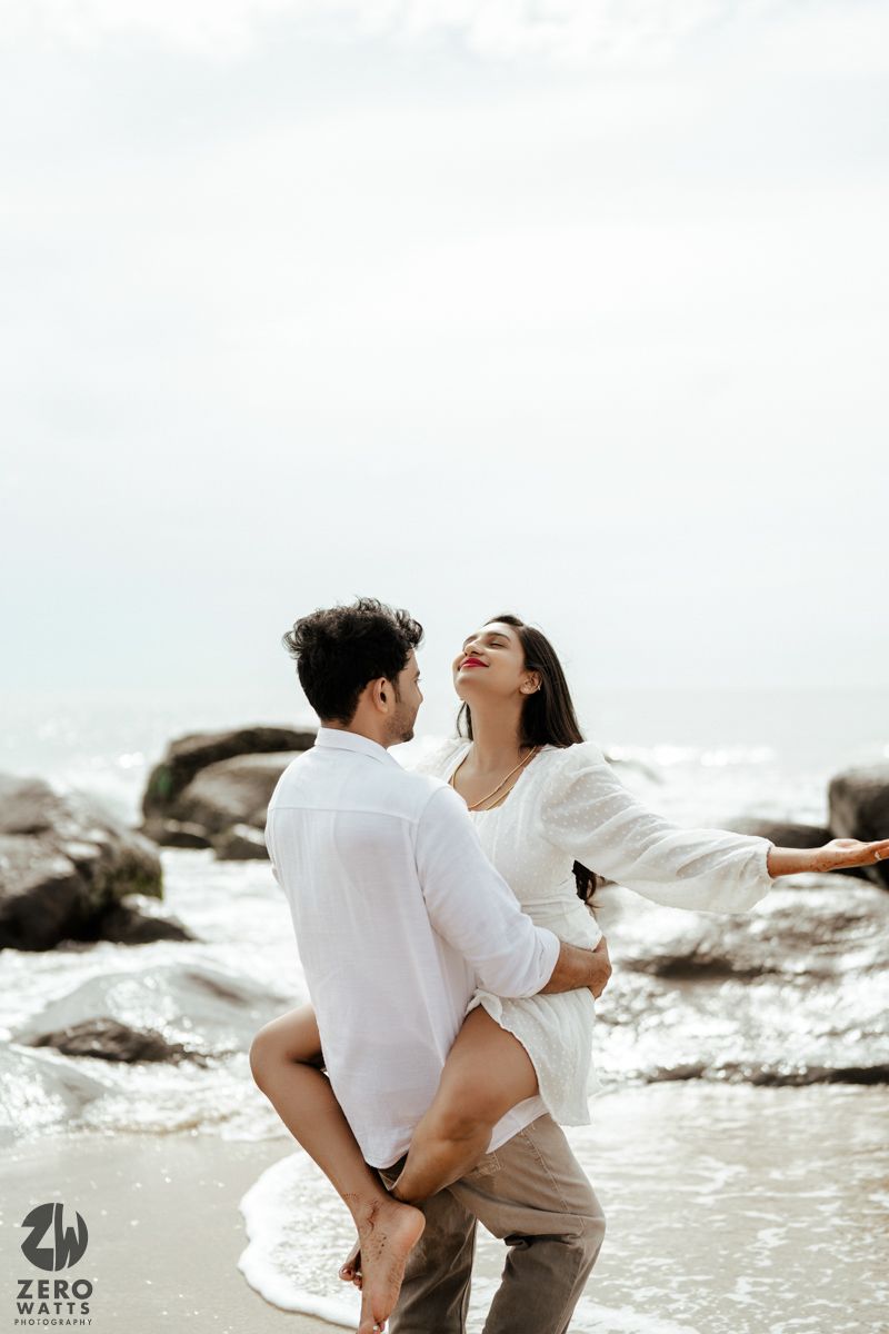 A romantic couple in white embraces while standing among coastal rocks with ocean waves.