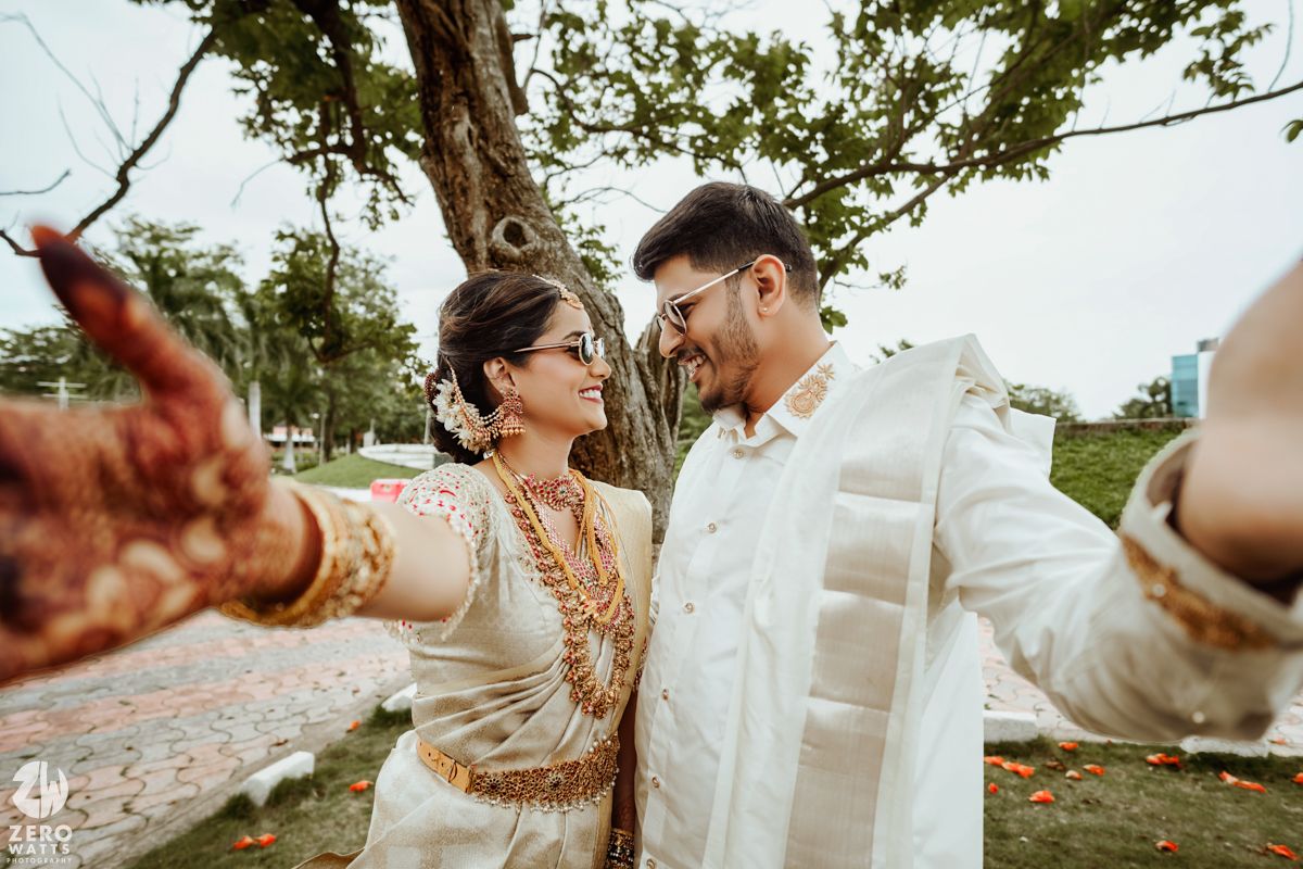 A couple in traditional dress taking a closeup selfie outdoors under a tree.