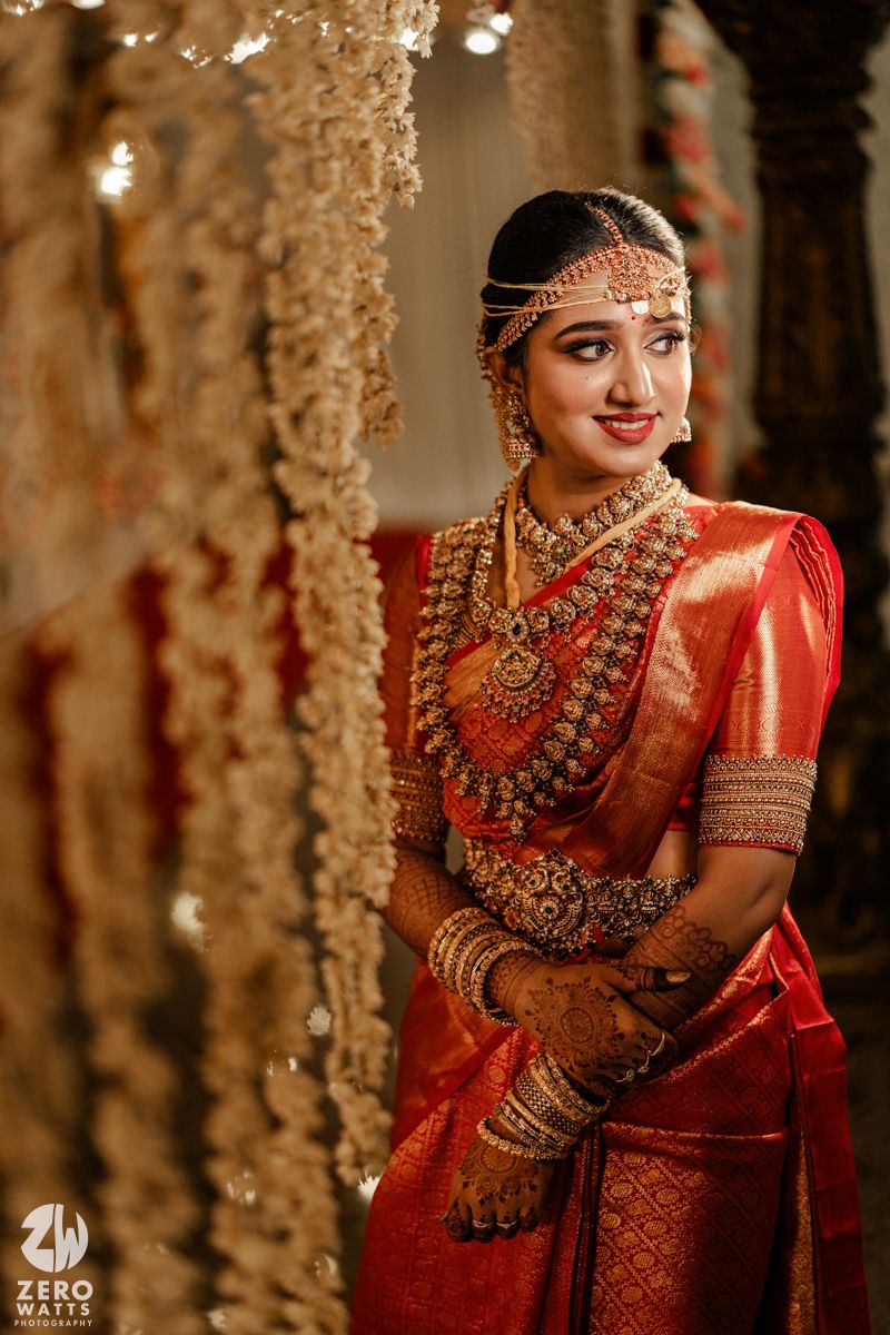 Traditional South Indian bride wearing luxurious red silk saree with ornate gold jewelry and headpiece amidst floral decor.