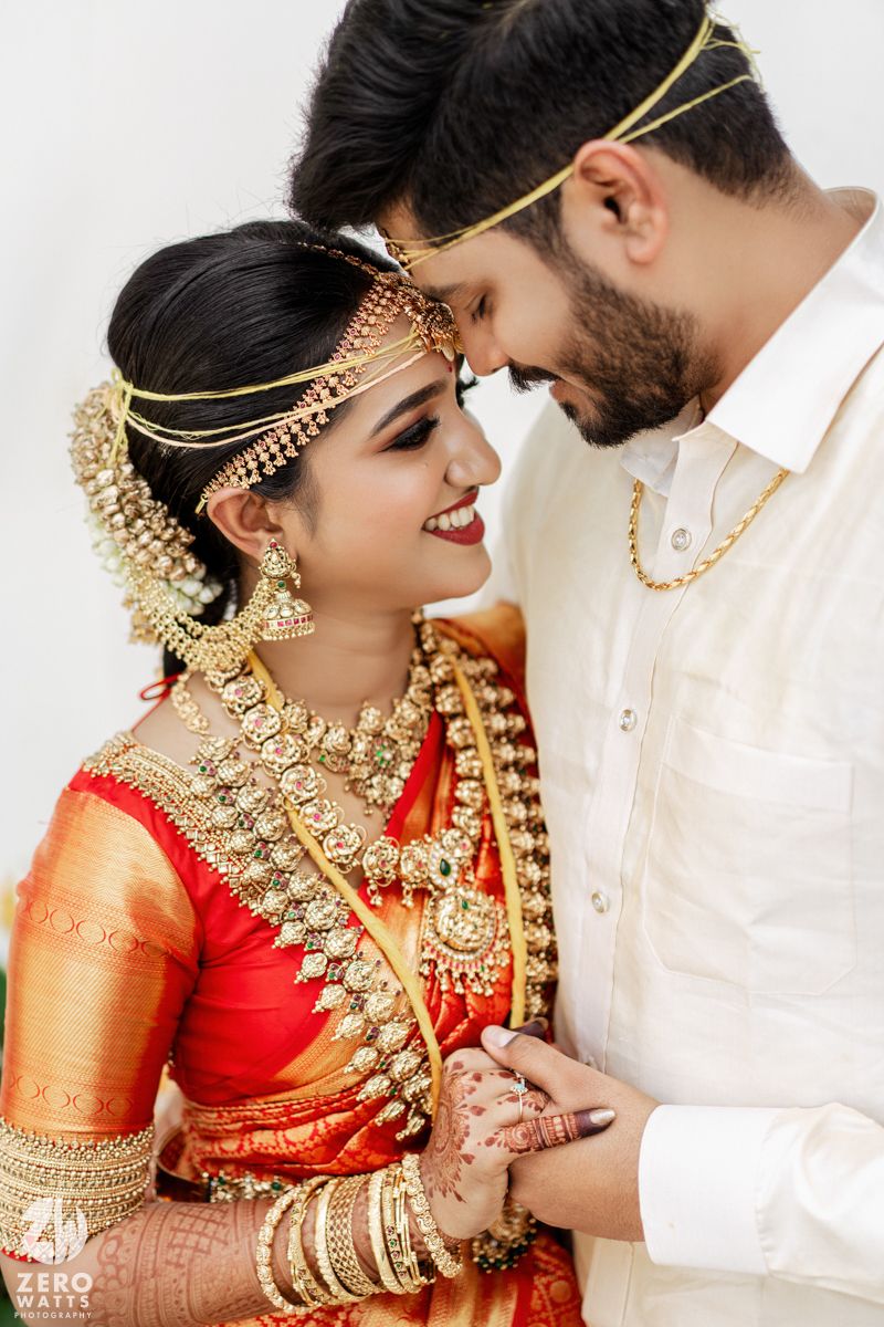 A couple in ornate Indian wedding attire share an intimate moment wearing traditional gold jewelry.