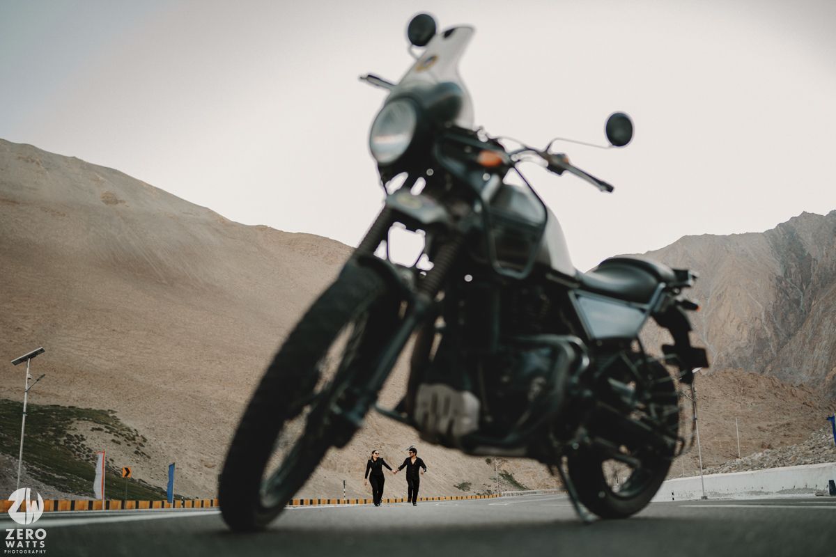 A motorcycle is silhouetted against rugged mountain terrain with two figures in the distance.