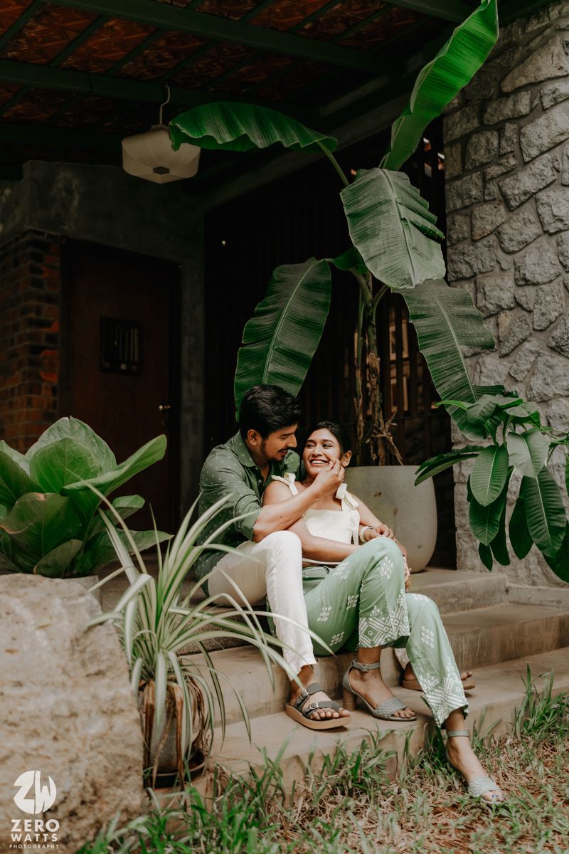 A romantic couple in white clothing shares playful moments on a stone wall amid tropical greenery.