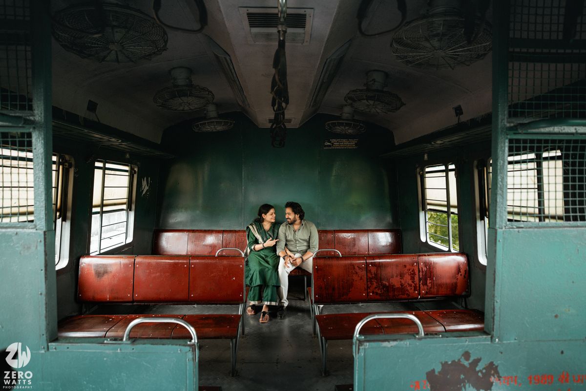 Two people sit close together on red seats inside an empty vintage train carriage with teal walls.