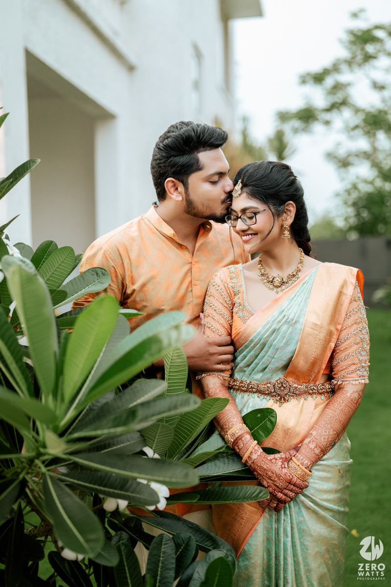 Romantic couple poses together in traditional Indian attire against lush tropical foliage and white columns.