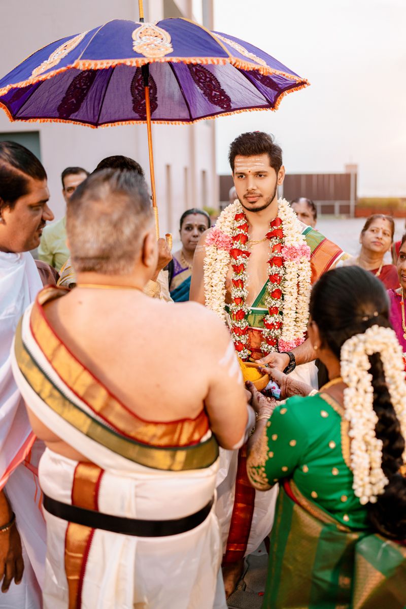 A traditional South Indian wedding ceremony under a decorative purple umbrella with guests and priests gathered around.