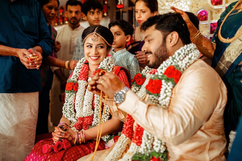 Traditional South Indian wedding ceremony with couple wearing red and cream garments adorned with flower garlands.